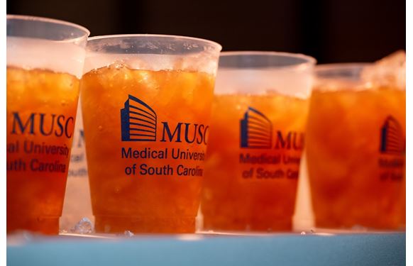 A row of plastic cups with the MUSC logo on them contain a brown-orange liquid. 