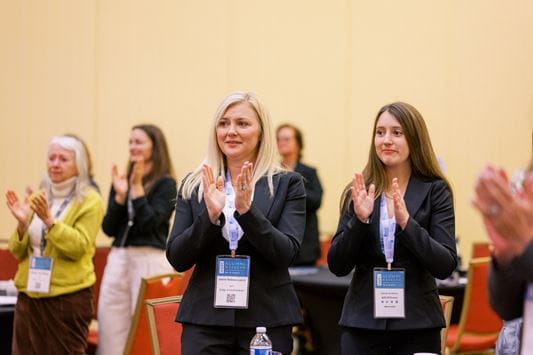 A group of 5-6 women dressed in casual business attire and wearing name tags stand to applaud. 