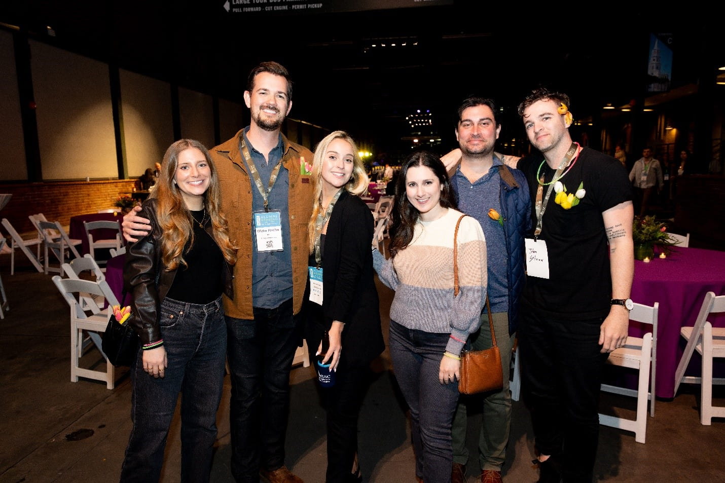 Group of 6 smiling people dressed in casual clothes and wearing name tag lanyards stand together with their arms around each other 