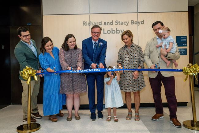 Six adults and two children stand behind a blue ribbon with the words “Grand Opening” on it. The adults (and one small girl) are all using blue scissors to cut the ribbon. On the wall behind them it sayd “Richard and Stacy Boyd Lobby” 