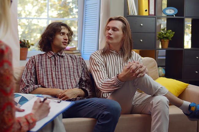Unhappy gay couple sitting on sofa and talking to psychologist. Portrait of homosexual male couple visiting family relationships expert discussing problems together