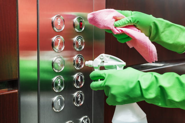 A person wearing green gloves uses a disinfectant spray and a wet wipe to clean an elevator button control panel