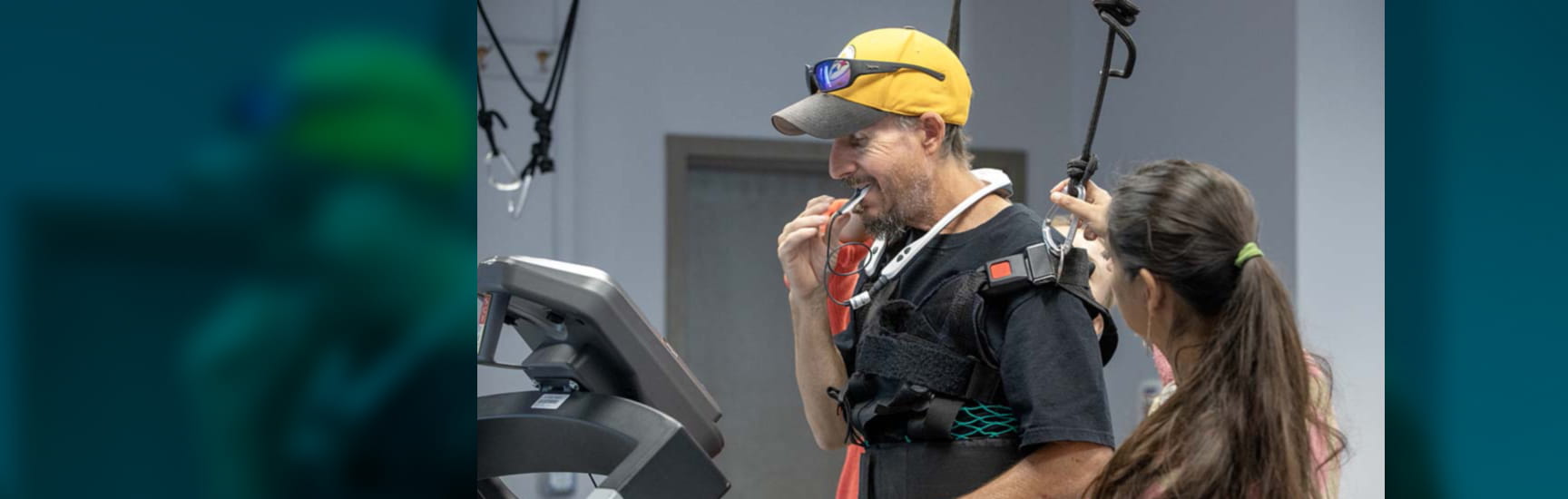 Man wearing a baseball cap puts a device in his mouth. A harness is helping to support him, as is a woman standing beside him.