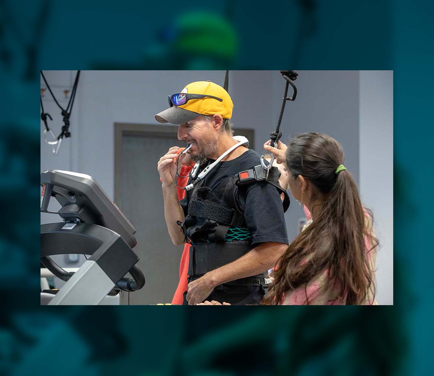 Man wearing a baseball cap puts a device in his mouth. A harness is helping to support him, as is a woman standing beside him. She has a long dark ponytail.
