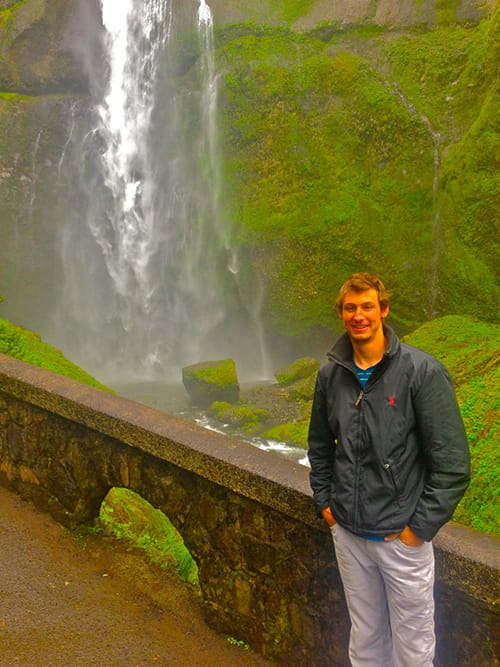 A young man stands in front of a low stone wall behind which is a lush green mountain and a waterfall