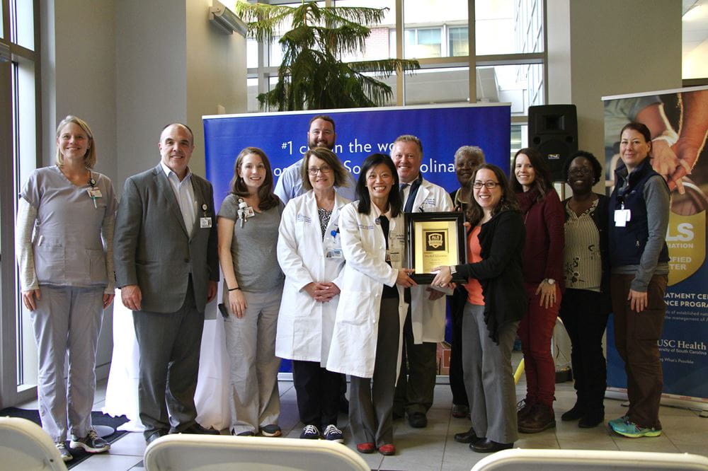 Members of MUSC's team as well as those from ALSA gather to pose for a photo holding a plaque commemorating MUSC being designated a Center of Excellence by the ALSA. 