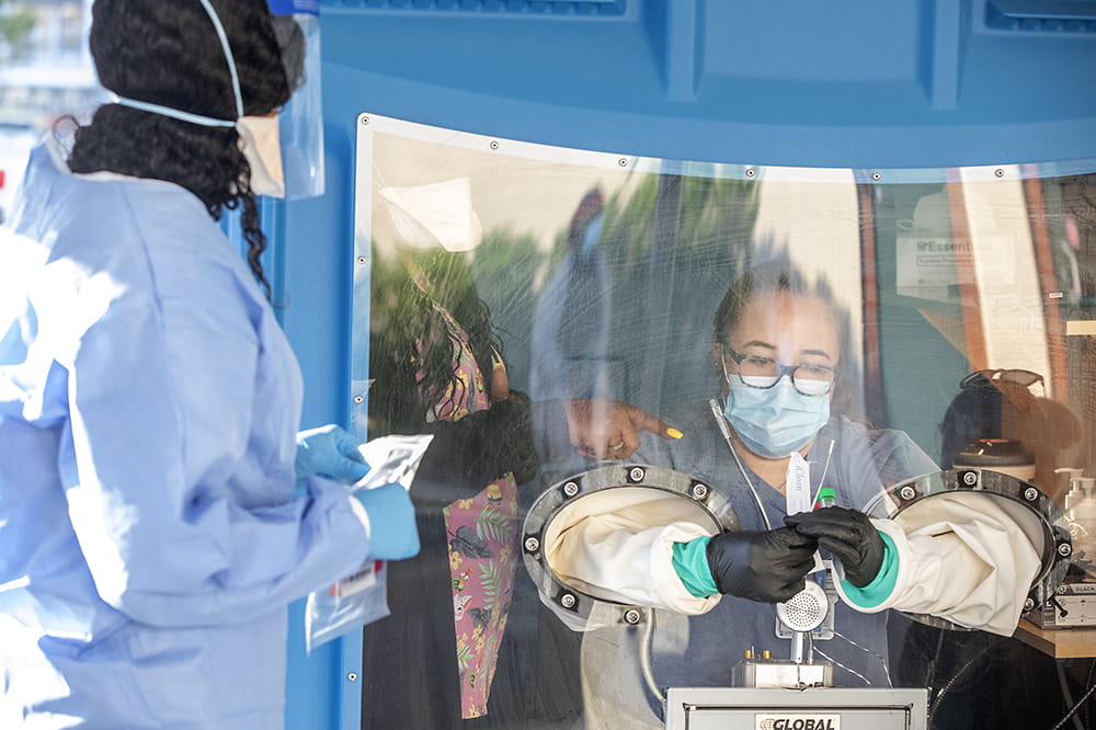 closeup of a masked woman in a booth putting her arms through gloves attached to holes in the wall while a woman inside the booth with her points what to do and a woman outside the booth in personal protective equipment watches