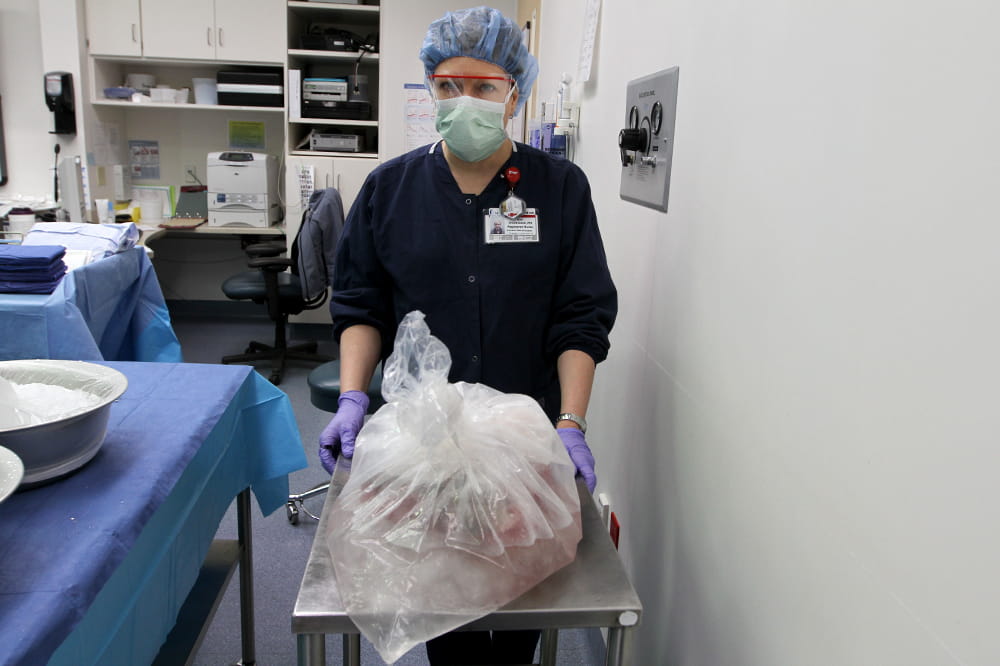 a nurse in scrubs wheels into the operating room a metal cart with a large semi clear plastic bag holding a large item that appears cold