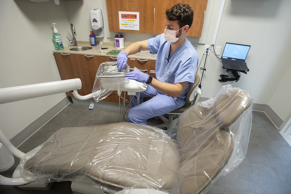 a man in blue scrubs opens a dental instrument kit in front of an empty exam chair