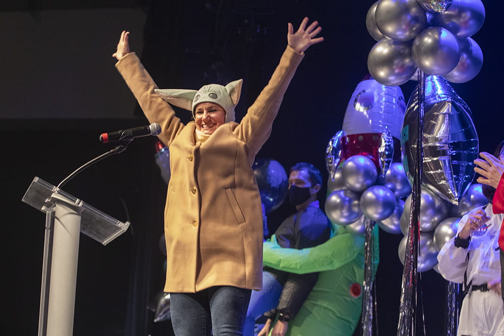 Sophia Sourlis, a woman in a khaki jacket and Yoda hat, puts her arms in the air on stage