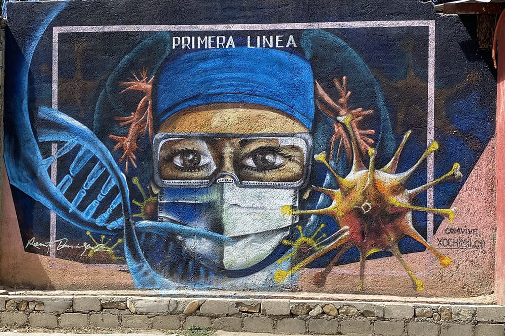 a street mural of a woman in a mask and surgical cap with a giant covid virus and DNA strand and the words "primera linea" or front line