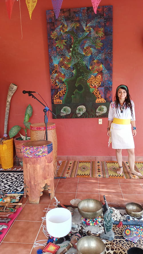 young woman standing in a colorful room of pink wall and terra cotta floor tile with traditional drums and microphone set up as for a ceremony