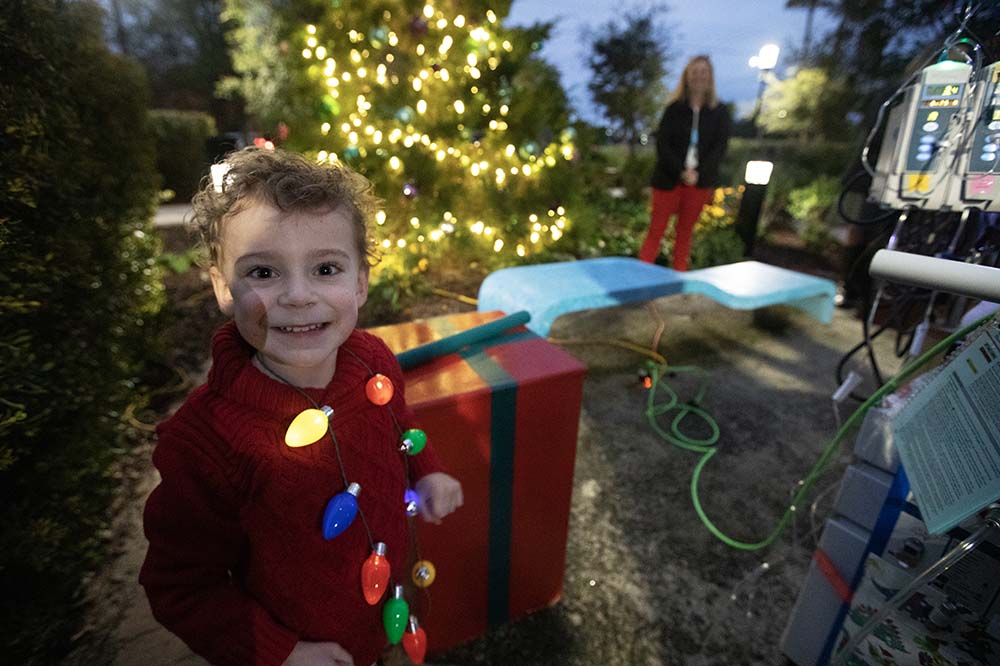 Boy in a red sweater wearing a Christmas light necklace smiles in front of the Angel Tree.