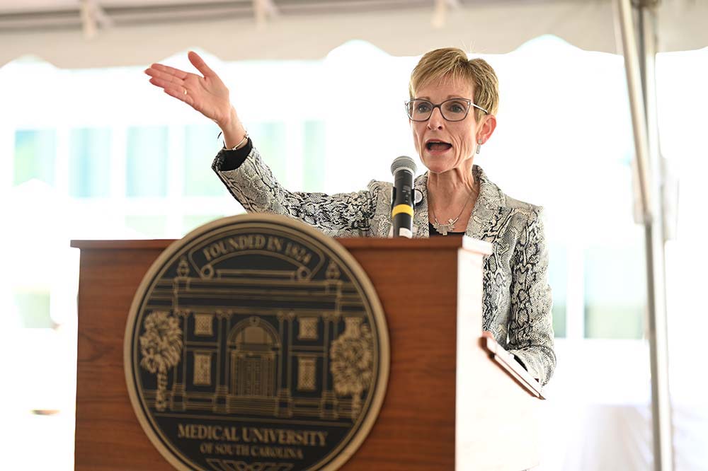 Woman wearing glasses stands behind a podium gesturing with her right hand.