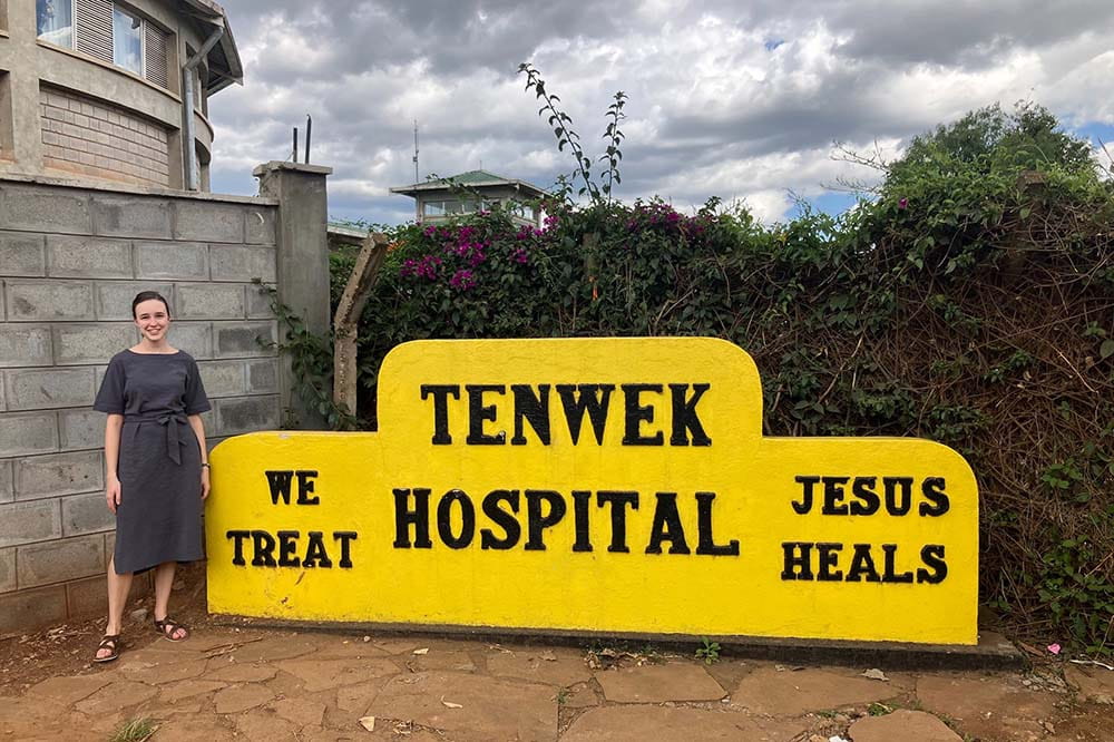 Woman wearing a dress stands beside a sign that says Tenwek Hospital. We treat. Jesus heals.