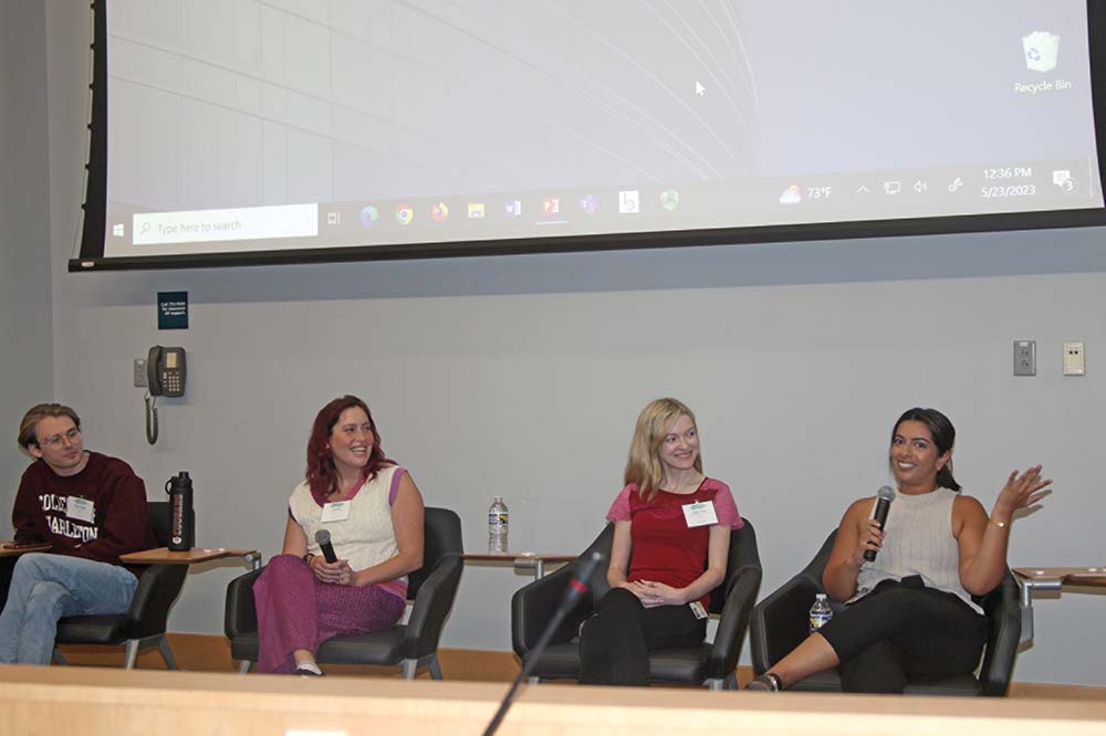 Four young women are seated facing an audience. The one on the far right is gesturing as she speaks.