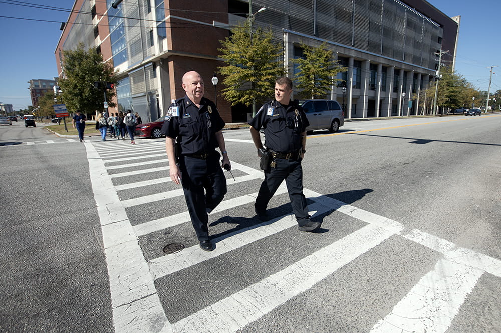 Two men in police uniforms walk in a crosswalk. There is a large building behind them.