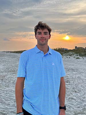 Young man in blue polo shirt stands on beach with sun behind him.