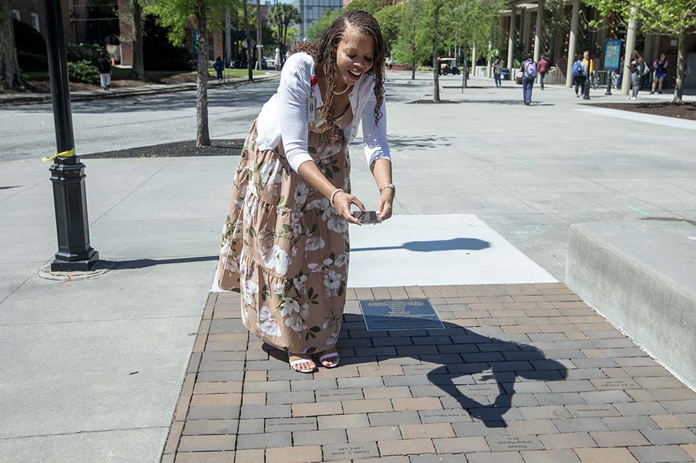 A woman wearing a dress leans forward to take a photo of a brick.