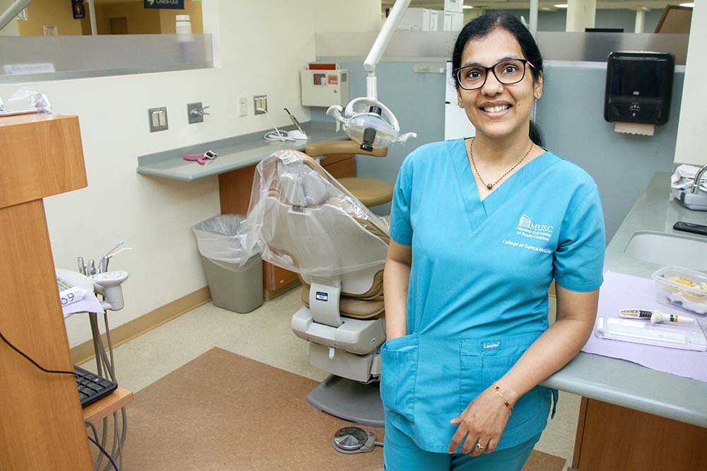 Women wearing blue scrubs and glasses smiles as she leans against a counter. Her dark hair is pulled back.