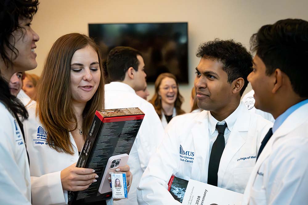 Four medical students wearing white coats talk in a crowded room.