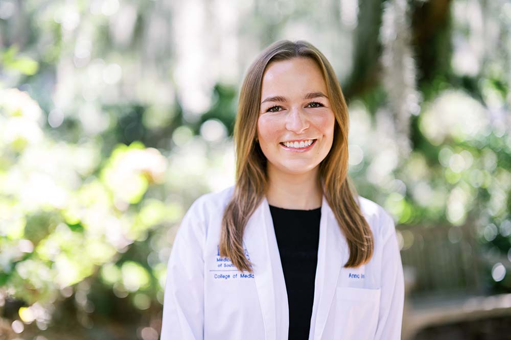 Young woman with light brown hair is wearing a white lab coat. She is standing outside.