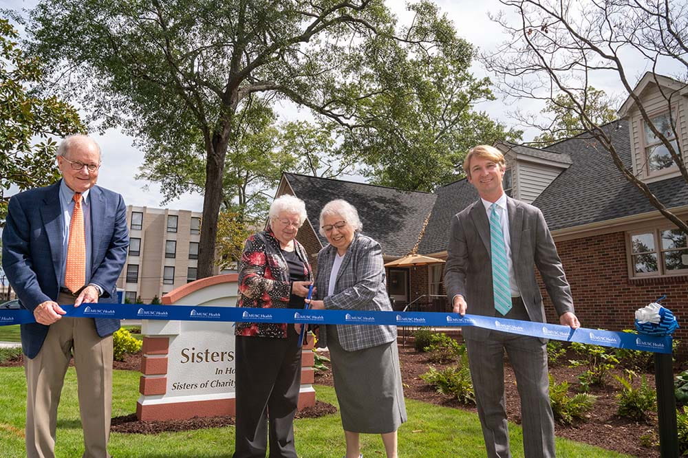 Two men and two women prepare to cut a large blue ribbon in front of a brick house.